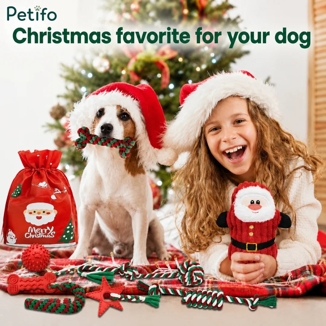 A Christmas scene showing a dog wearing a Santa hat holding a bone toy in its mouth next to a smiling young girl holding a plush Santa toy, with a Christmas tree and a red gift bag in the background.
