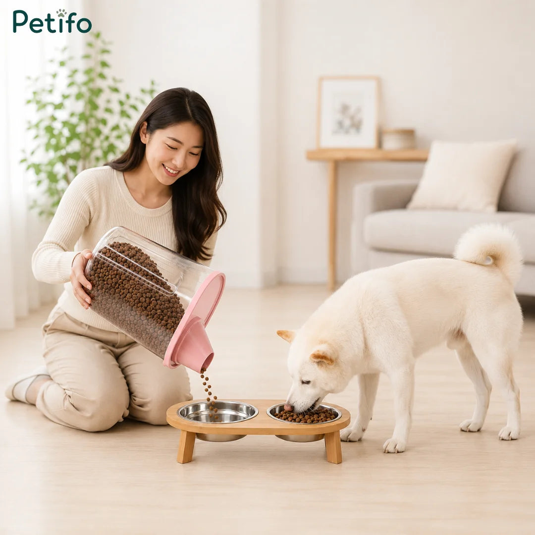 Young woman pouring dry kibble from a storage container into an elevated wooden double pet bowl as a white dog eats.