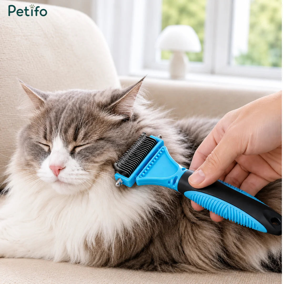 Petifo double-sided deshedding comb being gently used by a hand to brush the fur of a fluffy grey and white cat resting on a couch.