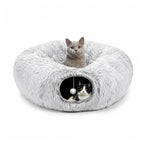A gray cat sitting inside a plush donut bed with a black and white cat peeking out of the tunnel entrance