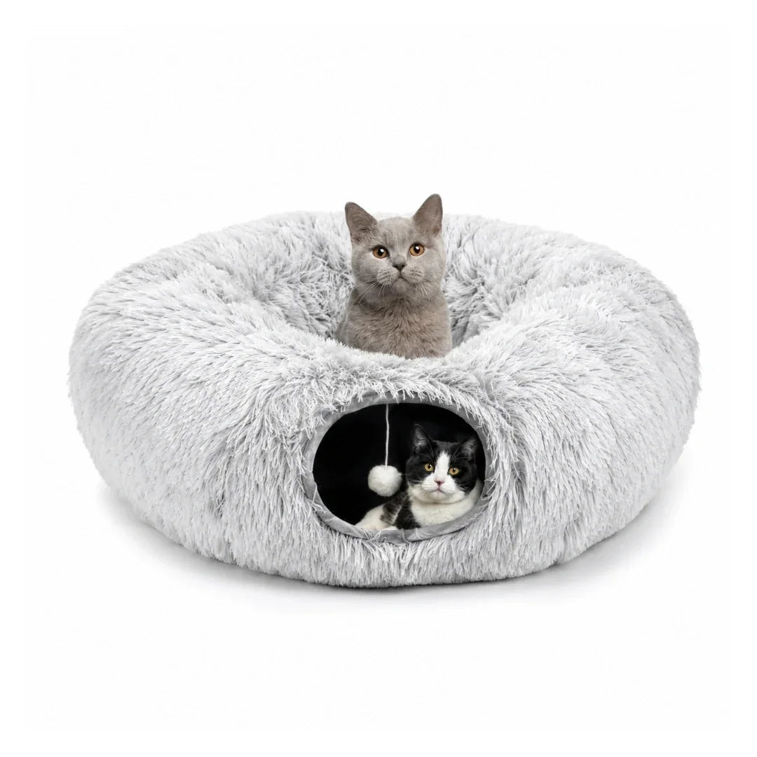 A gray cat sitting inside a plush donut bed with a black and white cat peeking out of the tunnel entrance