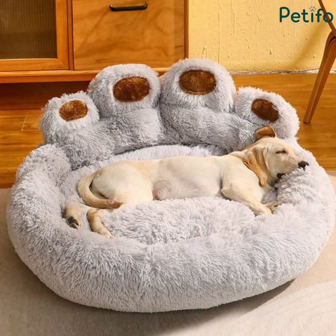 A dog sleeping soundly in a plush gray paw bed with brown paw pads on a wooden floor.